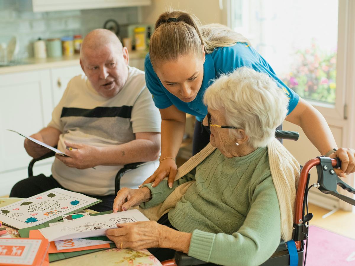 A carer leaning between two residents at a kitchen table, helping with an activity.
