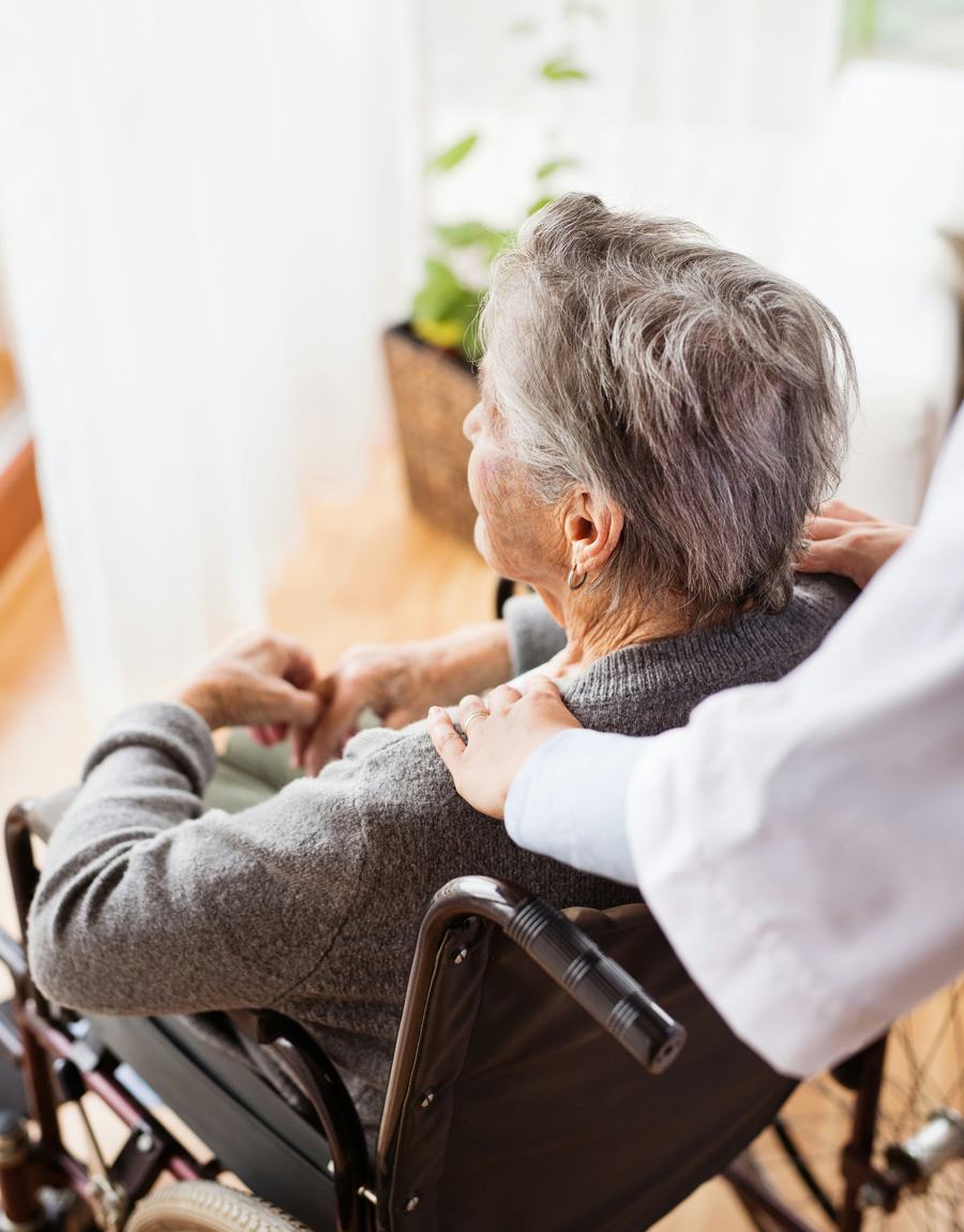 A resident in a wheelchair looking out of a sunlit window, a carer's hand resting on her shoulder.